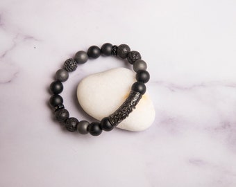 Peut inclure: Un bracelet en perles noires avec un charme argenté. Le bracelet est composé de pierres de lave noires, d'onyx noirs et de perles argentées. Le charme est un cylindre argenté avec un motif géométrique.
