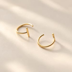 May include: A pair of gold-toned hoop earrings, each featuring a smooth, circular wire band that terminates in a perpendicular straight bar element. The earrings have a polished, reflective metallic finish and a simple, open-ended structural design.