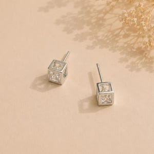 May include: A pair of silver-toned stud earrings featuring a hollow cube frame design. Each earring contains a clear, faceted square-cut stone suspended within the center of the metallic cube structure, complete with a thin post backing for attachment.