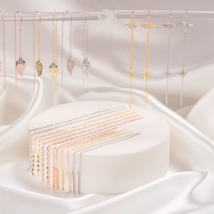 May include: Assortment of gold, silver, and rose gold dangle earrings with leaf, star, and bar designs. The earrings are displayed on a white circular stand.