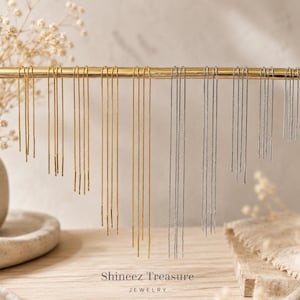 May include: A display of gold and silver threader earrings hanging from a gold bar. The earrings are of varying lengths and designs. The background is a neutral beige, with a vase of dried flowers on the left and the text "Shineez Treasure Jewelry" at the bottom.