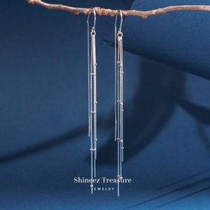 May include: Silver dangle earrings with multiple strands of chain and small beads hanging from a hook. The earrings are hanging from a branch against a blue background. The text "Shingez Treasure JEWELRY" is at the bottom of the image.