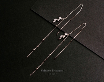 May include: A pair of long, delicate silver earrings with a leaf design and small clear crystals. The earrings are hanging on a black background.