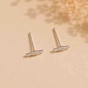 May include: A pair of silver-toned stud earrings featuring a horizontal bar design. Each earring consists of a straight post attached to a central bar, which is wrapped with a series of small, tightly coiled silver-toned wire rings.