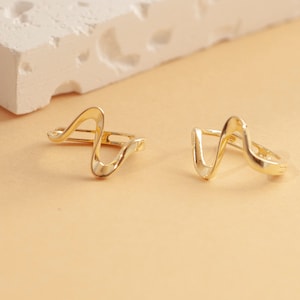 May include: A pair of gold-toned earrings with a wavy, abstract design. The earrings are made of metal and have a simple, modern style.