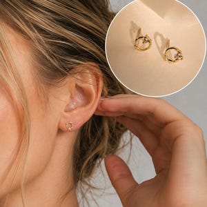 May include: A pair of gold stud earrings featuring a small, open circular design with a single clear gemstone accent. The earrings are shown worn in an earlobe and also displayed in a circular inset, highlighting the polished metal finish.