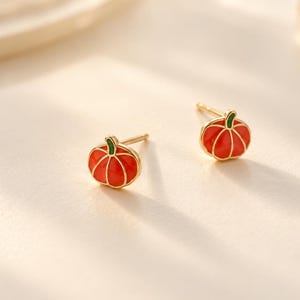 May include: A pair of stud earrings shaped like small pumpkins. Each earring features a bright orange enamel finish with gold-toned metal outlines and a small green stem detail at the top, complete with a standard post backing for secure wear.
