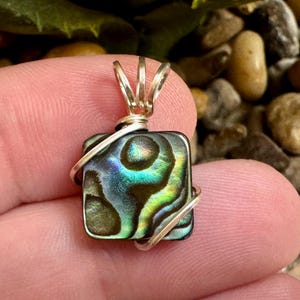 May include: Square pendant with iridescent abalone shell, wrapped in silver wire. The shell displays a mix of green, blue, and brown colors. The pendant has a silver bail for a necklace.