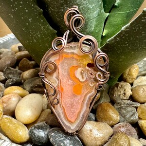 May include: A copper wire wrapped pendant featuring a natural agate stone with orange, white, and brown banding. The pendant is displayed on a bed of small stones.
