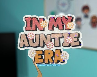 May include: A pink, orange, and white sticker with the text "In My Auntie Era" and small white flowers. The sticker is on a gold pair of tweezers.