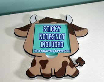 May include: A brown and tan cow-shaped sticky note holder with black trim. The holder has a rectangular opening for notes and features the text "STICKY NOTES NOT INCLUDED" in blue and white.