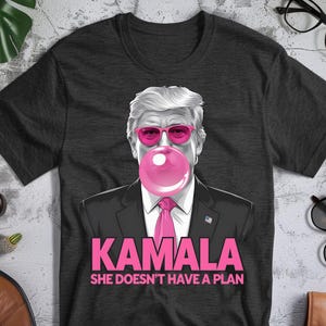 May include: Dark gray t-shirt featuring a graphic of a person with pink sunglasses blowing a bubblegum bubble. The text "KAMALA SHE DOESN'T HAVE A PLAN" is printed in pink below the graphic. The shirt is a crew neck.