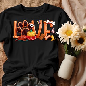 以下が含まれることがあります： オレンジ色の「LOVE」という文字が入った黒いTシャツ。足跡、犬、カボチャ、骨のグラフィックが特徴です。シャツには小さな蝶のグラフィックが含まれています。シャツの右側には花瓶に入った花があります。