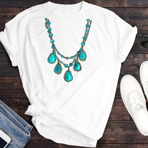 May include: White t-shirt featuring a turquoise and gold necklace with large turquoise stones. The shirt is laid out on a wooden surface with a pair of white trainers and sunglasses.