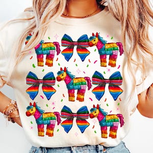 May include: Cream-colored t-shirt featuring a repeating pattern of colorful piñata horses and large rainbow bows. The piñatas are in shades of pink, blue, yellow, and green. Confetti and star accents are scattered around the design.