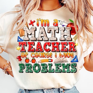 May include: Cream-colored t-shirt with a colorful graphic. The graphic reads "I'm a MATH TEACHER of course I have PROBLEMS" with math-themed illustrations like an apple, pencil, and open book. The text is in various colors.