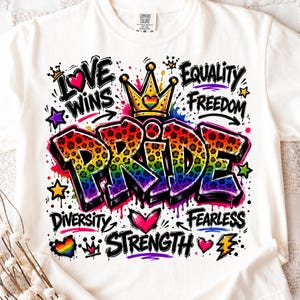 May include: A white t-shirt featuring a colourful graphic with the word PRIDE in a rainbow leopard print. The design includes a gold crown, hearts, stars, and the words LOVE WINS, EQUALITY, FREEDOM, DIVERSITY, FEARLESS, and STRENGTH in black text.