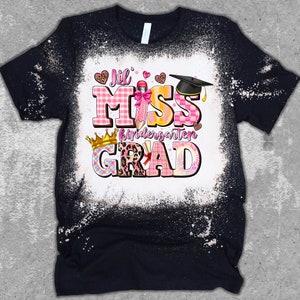 May include: Black t-shirt with a white bleached design. The shirt features the text "Lil Miss Kindergarten Grad" in pink, yellow, and plaid patterns, with a graduation cap and crown graphic.