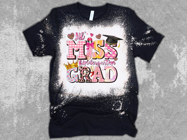 May include: Black t-shirt with a white bleached design. The shirt features the text "Lil Miss Kindergarten Grad" in pink, yellow, and plaid patterns, with a graduation cap and crown graphic.