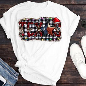 K&ouml;nnte beinhalten: Wei&szlig;es T-Shirt mit einem Grafikdruck des Wortes "TEXAS" in rot-schwarz-wei&szlig;em Karomuster mit einem rot-wei&szlig;en Stern und einer Weihnachtsm&uuml;tze. Die Grafik ist von einem Leopardenmuster-Rand und wei&szlig;en Lichtern umgeben.