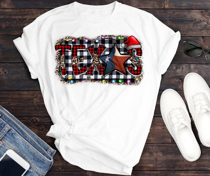 K&ouml;nnte beinhalten: Wei&szlig;es T-Shirt mit einem Grafikdruck des Wortes "TEXAS" in rot-schwarz-wei&szlig;em Karomuster mit einem rot-wei&szlig;en Stern und einer Weihnachtsm&uuml;tze. Die Grafik ist von einem Leopardenmuster-Rand und wei&szlig;en Lichtern umgeben.