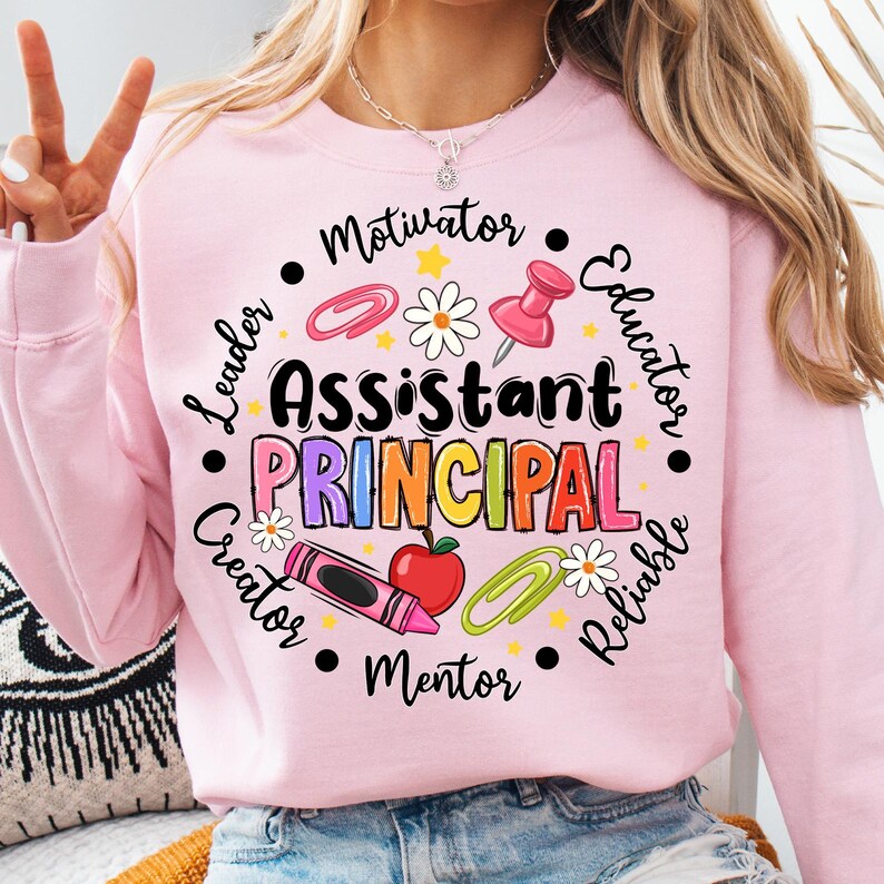 May include: A light pink sweatshirt with the words "Assistant Principal" in colorful letters. The design includes school-themed graphics like a red apple, crayon, paperclip, and push pin. The words "Motivator", "Educator", "Reliable", "Mentor", "Creator", and "Leader" are also included.