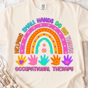 May include: Cream-colored t-shirt with a colorful rainbow design and the text "Helping Small Hands Do Big Things." The design includes handprints and the words "Occupational Therapy."