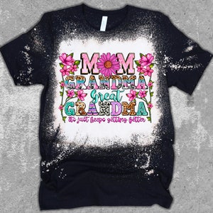 May include: Black t-shirt with a bleached white center. The shirt features the words "MOM GRANDMA GREAT GRANDMA" in colorful, floral-decorated text. The phrase "It just keeps getting better" is below the text.