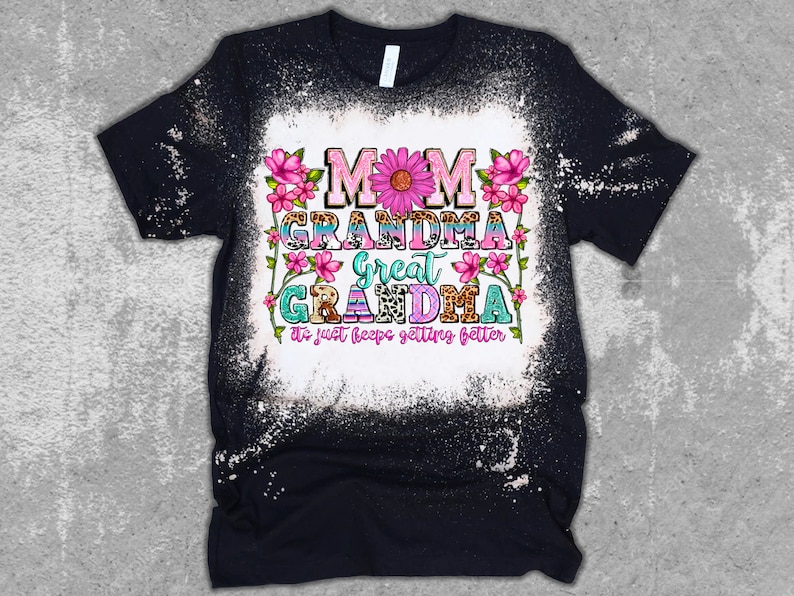 May include: Black t-shirt with a bleached white center. The shirt features the words "MOM GRANDMA GREAT GRANDMA" in colorful, floral-decorated text. The phrase "It just keeps getting better" is below the text.