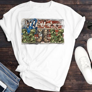 May include: White t-shirt featuring the text "Military Mom" in a patriotic design. The design incorporates the American flag, camouflage lettering, and military boots. Sunglasses, white shoes, and jeans are also visible.