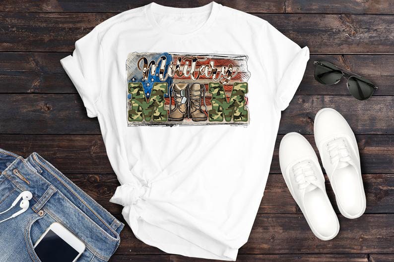 May include: White t-shirt featuring the text "Military Mom" in a patriotic design. The design incorporates the American flag, camouflage lettering, and military boots. Sunglasses, white shoes, and jeans are also visible.
