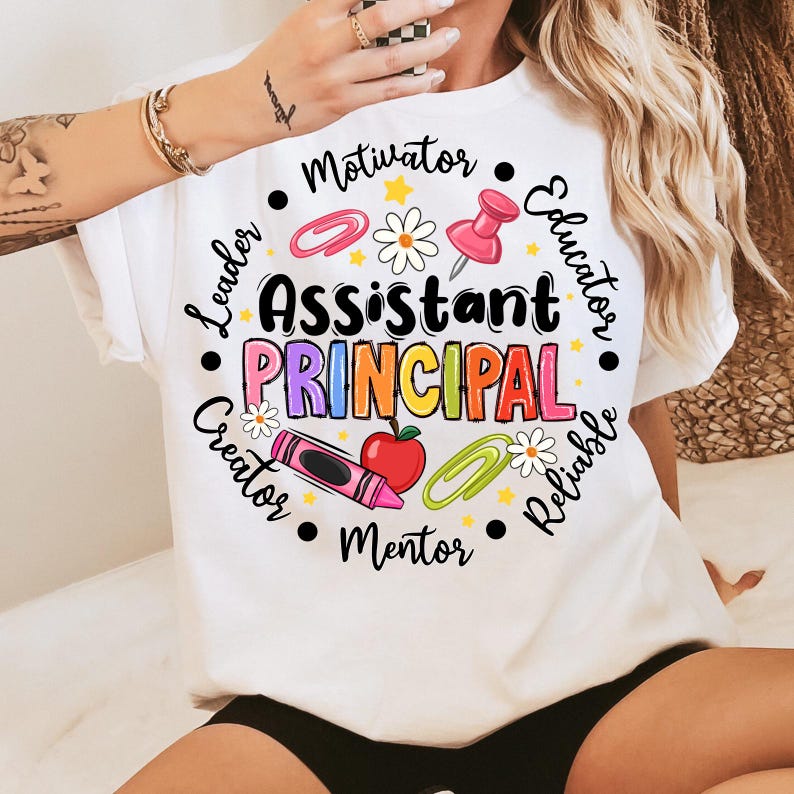 May include: White t-shirt with the words "Assistant Principal" in colorful letters, surrounded by words like "Motivator" and "Educator." The design includes a push pin, paper clips, a crayon, an apple, and daisies.
