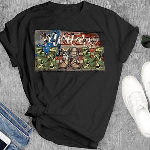 May include: Black t-shirt with the words "Military Mom" in a patriotic design. The word "MOM" is in camouflage print, and the shirt features an American flag and military boots graphic. Sunglasses, white shoes, and jeans are also visible.