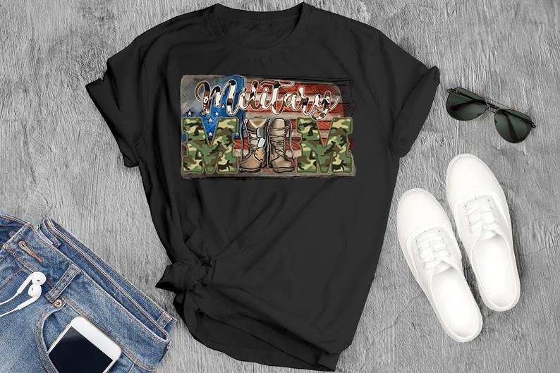 May include: Black t-shirt with the words "Military Mom" in a patriotic design. The word "MOM" is in camouflage print, and the shirt features an American flag and military boots graphic. Sunglasses, white shoes, and jeans are also visible.