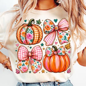Puede incluir: Una camiseta de color crema con un estampado gráfico de dos calabazas y dos lazos. Una calabaza tiene un patrón de cuadros naranja, mientras que la otra es naranja sólido. Ambas están decoradas con motivos florales, rayas rosas y pequeños corazones y estrellas.