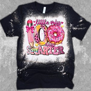 May include: A black t-shirt with a bleach-splattered design featuring a pink pencil, a donut, and the text "little miss 100 days smarter".