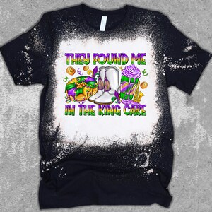 May include: A black t-shirt with a white bleach design. The design features a king cake, a pair of white boots, and the text "They found me in the king cake".