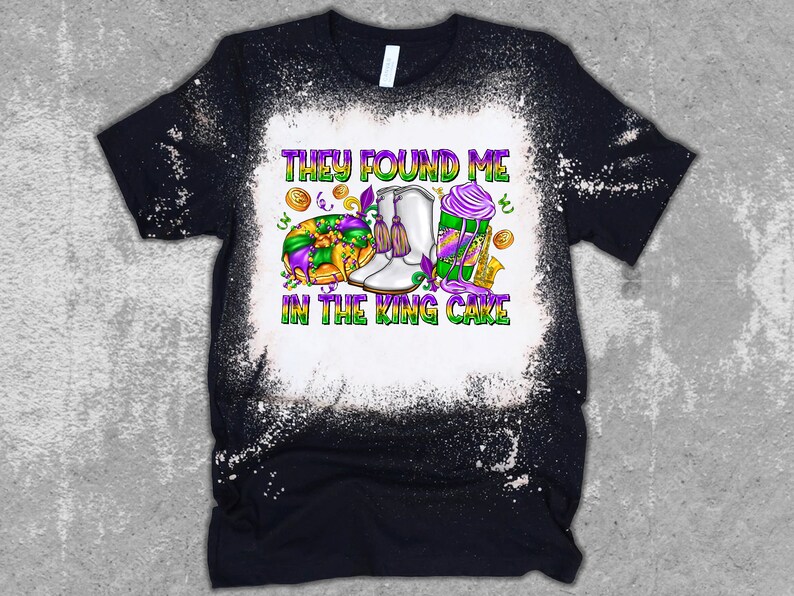 May include: A black t-shirt with a white bleach design. The design features a king cake, a pair of white boots, and the text "They found me in the king cake".