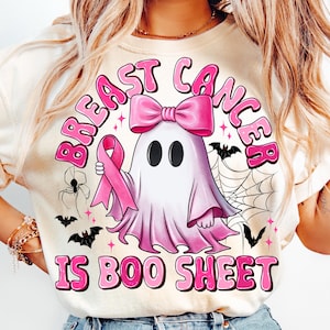 May include: A cream-colored t-shirt featuring a graphic of a white ghost wearing a pink bow and holding a pink awareness ribbon. The text reads BREAST CANCER IS BOO SHEET in pink letters, surrounded by black spiderwebs, spiders, and bats.