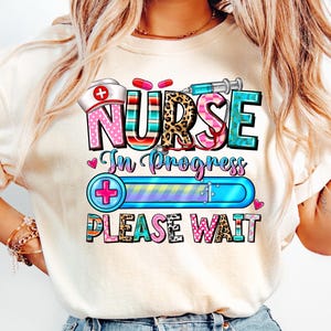 May include: A cream-coloured t-shirt with a vibrant graphic. The design includes the word "NURSE" in diverse patterns, a nurse's hat, a syringe, and the phrases "In Progress" and "PLEASE WAIT".
