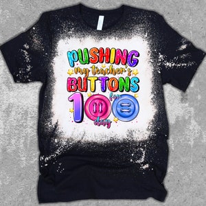 K&ouml;nnte beinhalten: Ein schwarzes T-Shirt mit einem gebleichten Design, auf dem "Pushing my teacher's buttons for 100 days" mit bunten Kn&ouml;pfen und Sternen steht.