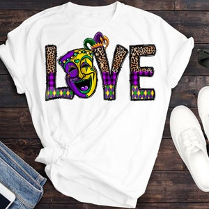 May include: White t-shirt with a purple, green, and gold "LOVE" graphic featuring a jester mask. The letters are decorated with leopard print and plaid patterns.