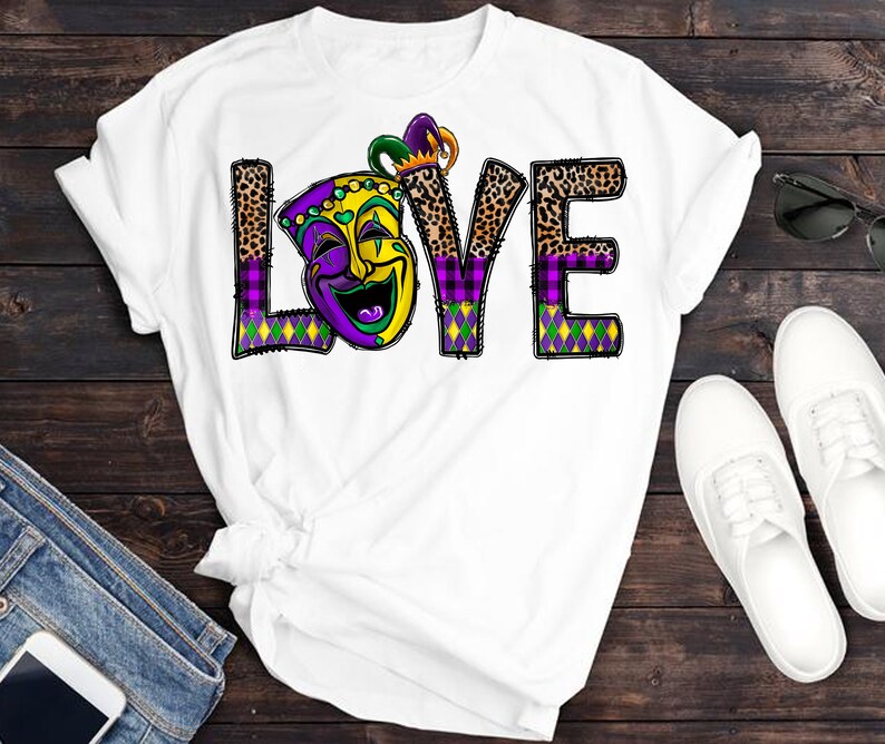 May include: White t-shirt with a purple, green, and gold "LOVE" graphic featuring a jester mask. The letters are decorated with leopard print and plaid patterns.