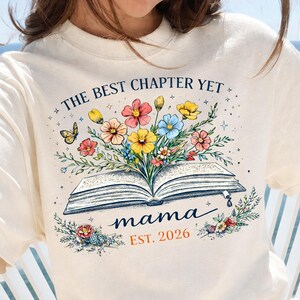 May include: Cream-colored sweatshirt with a floral book design. The design features an open book with colorful flowers, a butterfly, and the text "THE BEST CHAPTER YET" and "mama EST. 2026".