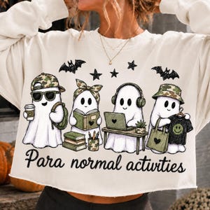 Peut inclure: Un sweat-shirt court de couleur crème avec un motif représentant quatre fantômes occupés à diverses activités. Les fantômes portent des chapeaux, utilisent un ordinateur portable, lisent des livres et tiennent des objets. Le texte "Para normal activities" est imprimé en dessous.