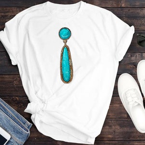 May include: A white t-shirt featuring a turquoise gemstone earring pendant. The earring is a teardrop shape with a round stone at the top. The t-shirt is displayed on a wooden surface.