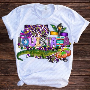 May include: White t-shirt with a colorful graphic design featuring the word "LOUISIANA" in a purple, green, and gold font. The graphic also includes a green alligator, a purple and gold fleur-de-lis, and a Mardi Gras mask.