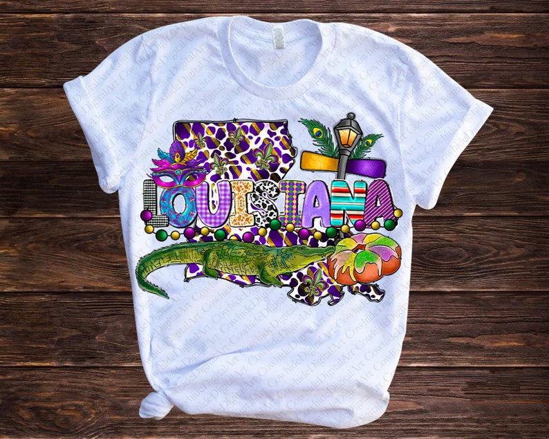 May include: White t-shirt with a colorful graphic design featuring the word "LOUISIANA" in a purple, green, and gold font. The graphic also includes a green alligator, a purple and gold fleur-de-lis, and a Mardi Gras mask.