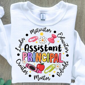 May include: White sweatshirt with the words "Assistant Principal" in colorful letters. The design includes a crayon, apple, paperclip, push pin, and daisy. Surrounding the words are the words "Leader, Motivator, Educator, Reliable, Creator, Mentor."