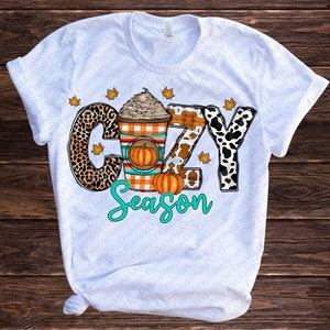 May include: White t-shirt with a graphic design of a coffee cup with whipped cream and a pumpkin, surrounded by animal print letters that spell out "Cozy Season".